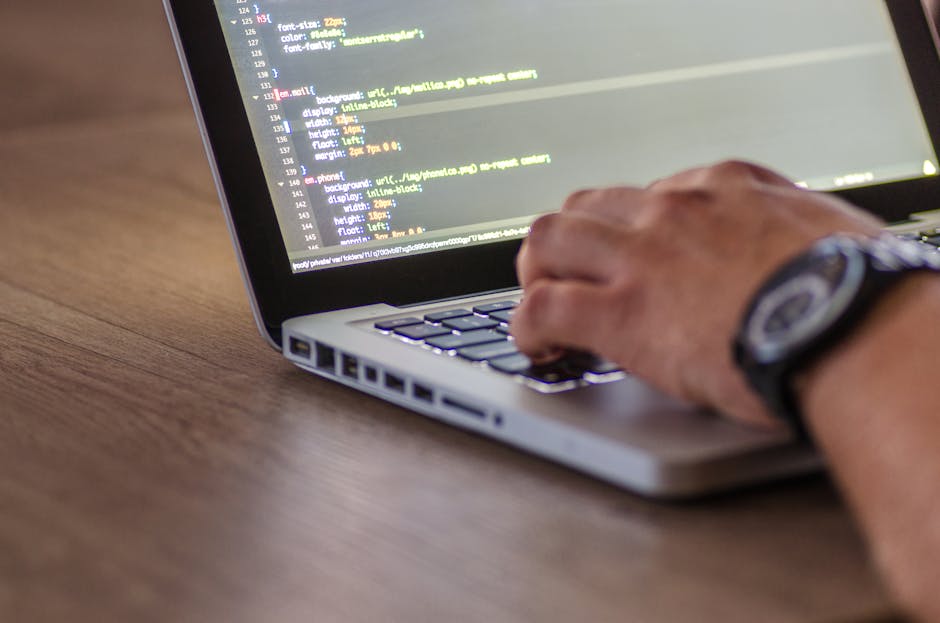 A close-up of a person's hand operating a laptop computer placed on a wooden desk, with the screen displaying lines of code or programming text. The person is wearing a wristwatch on their left wrist 