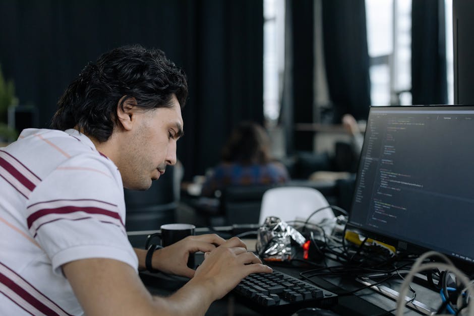 A close-up of a person's hand operating a laptop computer placed on a wooden desk, with the screen displaying lines of code or programming text. The person is wearing a wristwatch on their left wrist 